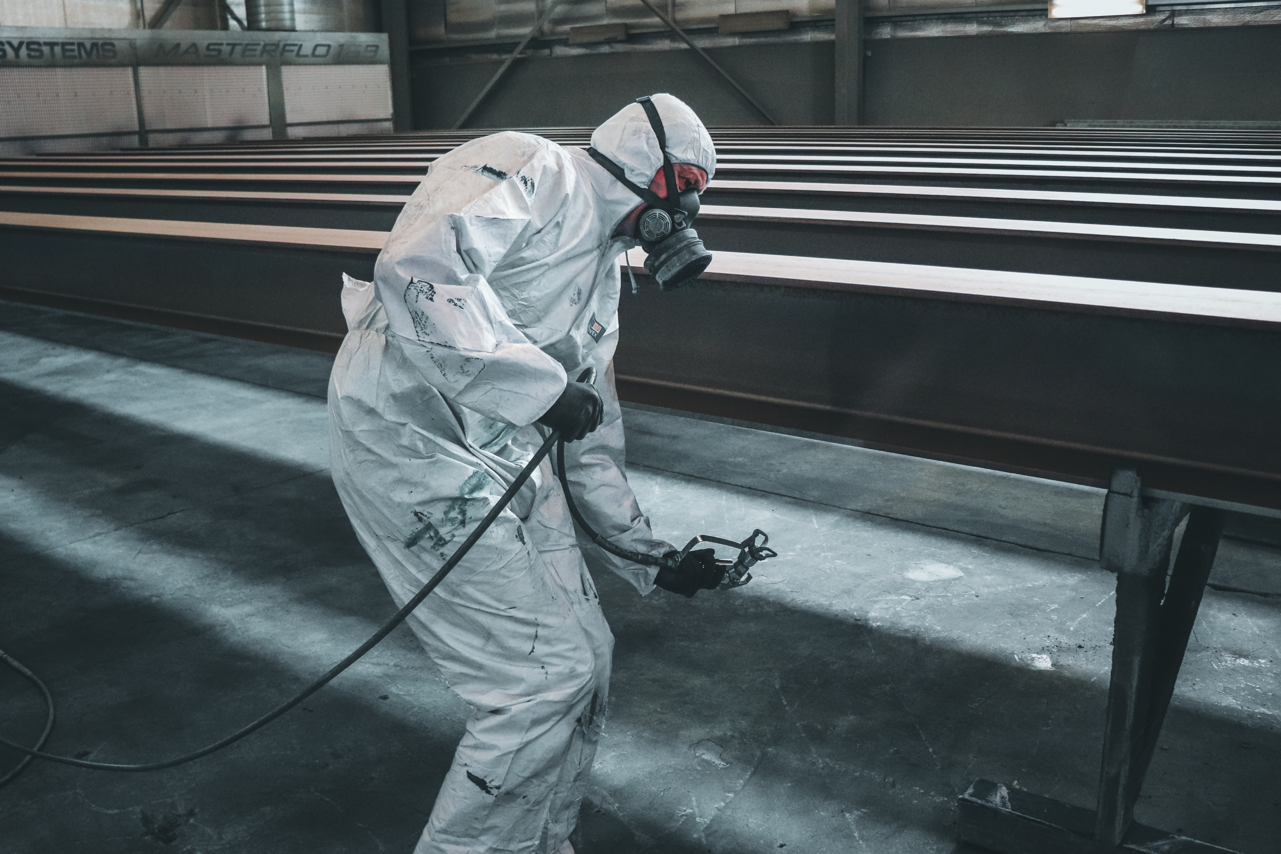 Worker in protective gear spray painting metal beams inside a factory setting.